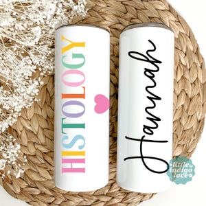 May include: Two white insulated tumblers. One features the word "HISTOLOGY" in rainbow colors with a pink heart. The other has the name "Hannah" in black script. Both tumblers have a silver rim.