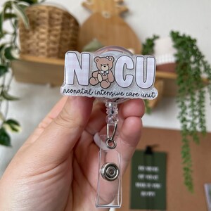 May include: A clear badge reel with a rectangular plastic clip and a retractable cord. The badge features the letters "NICU" in a white, block font over a pink and white gingham pattern. A teddy bear graphic is in the center, and the words "neonatal intensive care unit" are below.
