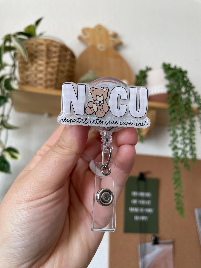 May include: A clear badge reel with a rectangular plastic clip and a retractable cord. The badge features the letters "NICU" in a white, block font over a pink and white gingham pattern. A teddy bear graphic is in the center, and the words "neonatal intensive care unit" are below.