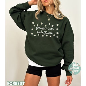 May include: A dark green sweatshirt with the text "physician assistant" in white lettering surrounded by white daisy flowers.