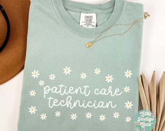 May include: A light blue t-shirt with the text "patient care technician" in white cursive lettering. The text is surrounded by white daisies with yellow centers. The shirt is a comfort colors brand.