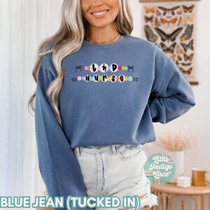 K&ouml;nnte beinhalten: Ein blaues Sweatshirt mit dem Text "L&D Nurse" in schwarzen Buchstaben, umgeben von bunten Perlen. Das Sweatshirt ist in eine blaue Jeans gesteckt.