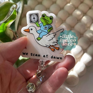 May include: A white acrylic badge reel featuring a green frog wearing blue scrubs and holding a CT scan image while riding a white goose with orange feet. The text "we scan at dawn" is printed in black below the illustration on the badge reel.