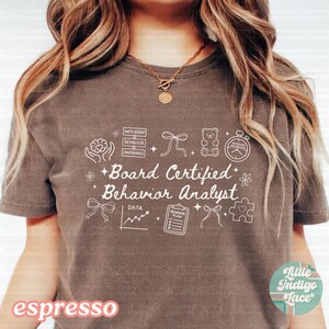 May include: Espresso-colored t-shirt with the text "Board Certified Behavior Analyst" in white, along with related graphic icons. The shirt features a crew neck and short sleeves. The words "little indigo lace" are also visible.