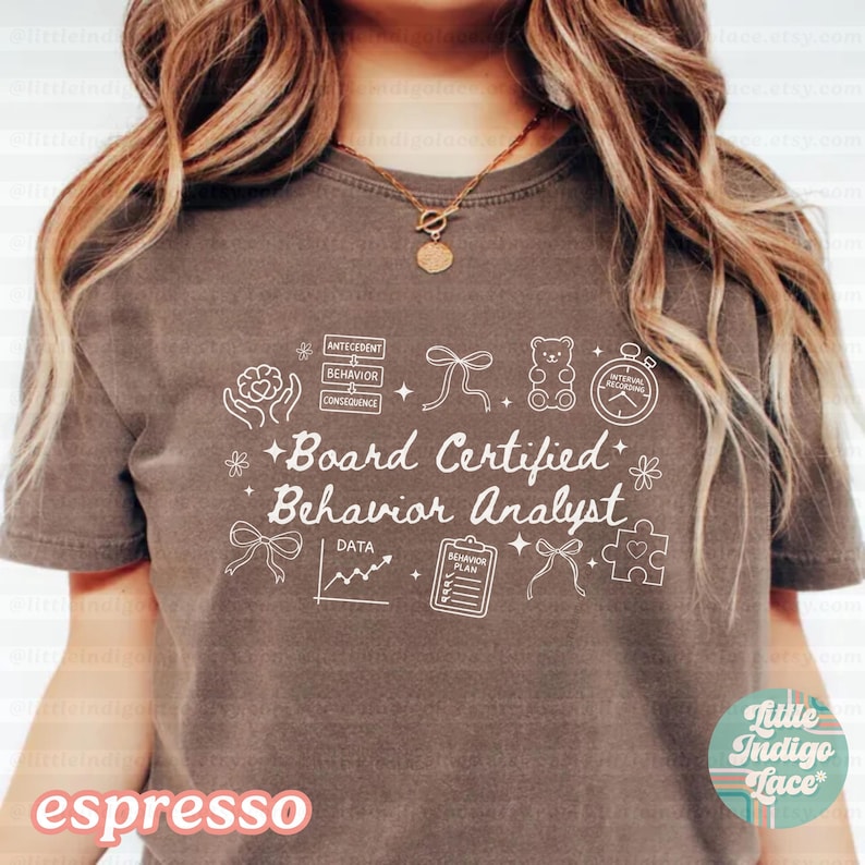 May include: Espresso-colored t-shirt with the text "Board Certified Behavior Analyst" in white, along with related graphic icons. The shirt features a crew neck and short sleeves. The words "little indigo lace" are also visible.