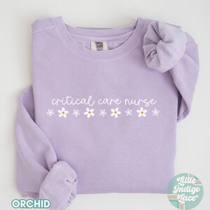 May include: A lavender sweatshirt with the text "critical care nurse" in white lettering and a row of white daisies below the text. The sweatshirt is folded and laid flat on a white surface.