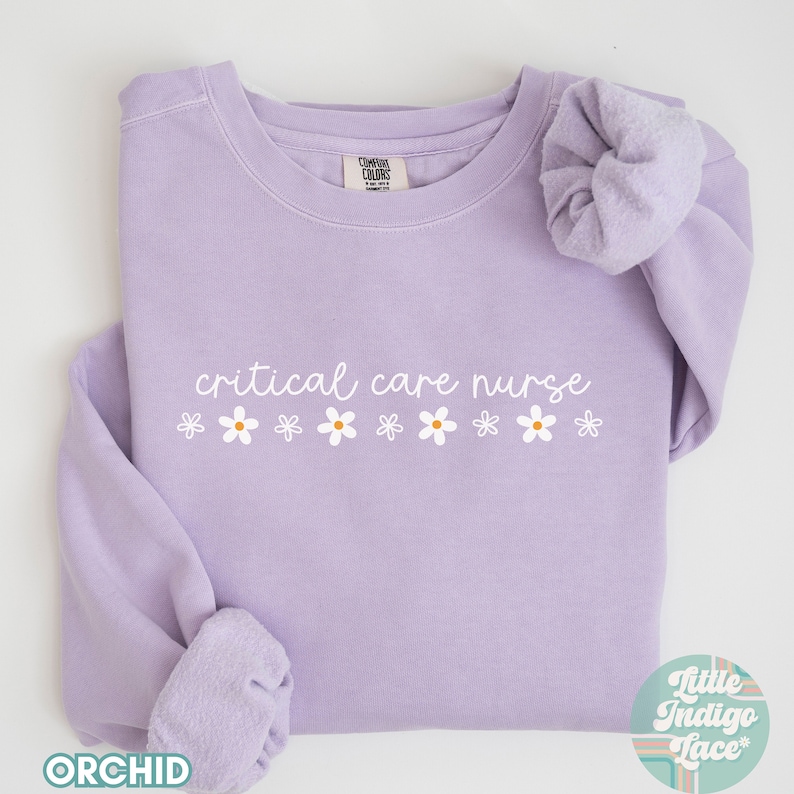 May include: A lavender sweatshirt with the text "critical care nurse" in white lettering and a row of white daisies below the text. The sweatshirt is folded and laid flat on a white surface.