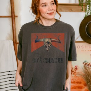 May include: A dark gray t-shirt with a vintage-style graphic of a bull skull with an American flag design. The text "90's COUNTRY" is printed below the graphic.
