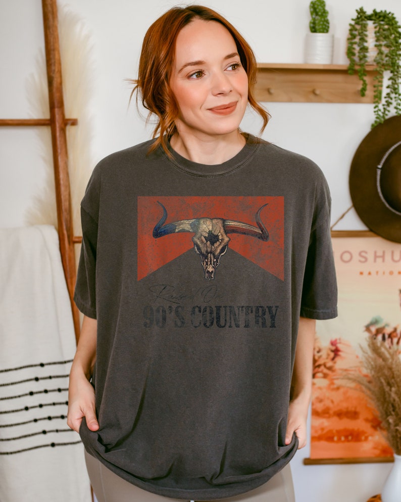 May include: A dark gray t-shirt with a vintage-style graphic of a bull skull with an American flag design. The text "90's COUNTRY" is printed below the graphic.