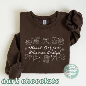 May include: A dark chocolate-colored sweatshirt with white text and graphics. The text reads "Board Certified Behavior Analyst" with related icons. The bottom of the sweatshirt has the words "dark chocolate" in a green font.