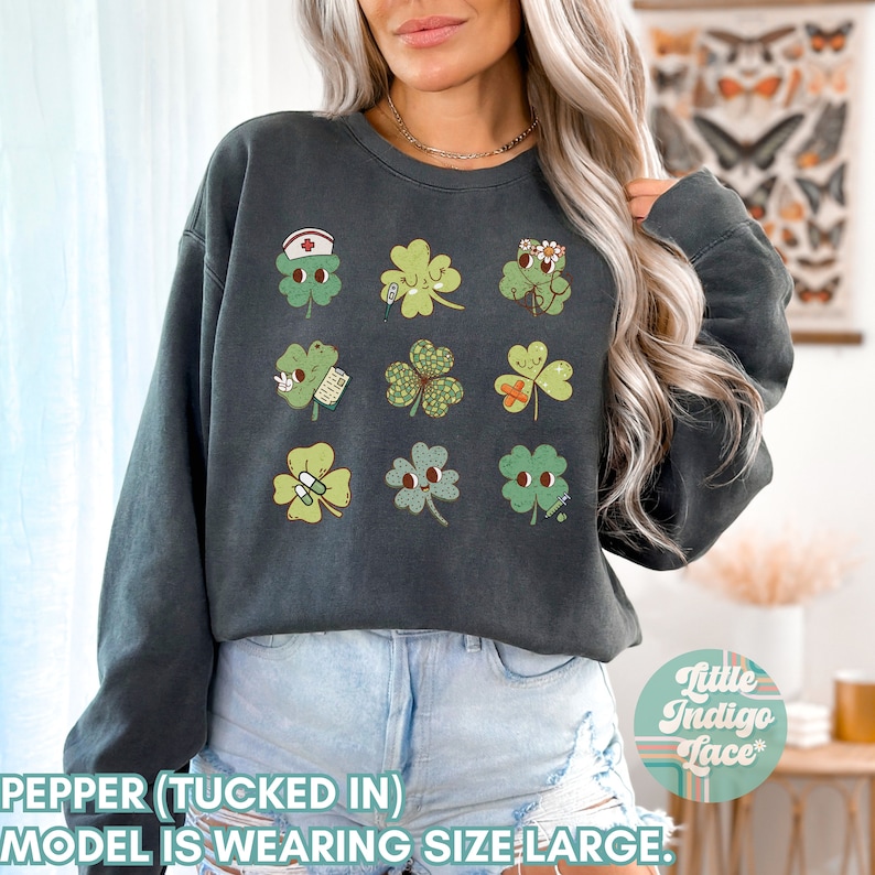 K&ouml;nnte beinhalten: Ein dunkelgraues Sweatshirt mit einem grafischen Druck von neun gr&uuml;nen vierbl&auml;ttrigen Kleebl&auml;ttern, jedes mit einem anderen Gesichtsausdruck und Detail. Die Kleebl&auml;tter beinhalten Designs wie eine Krankenschwesterm&uuml;tze, eine Blumenkrone, einen Verband und eine Pillenflasche. Der Text "Little Indigo Lace" ist in der unteren rechten Ecke des Bildes sichtbar.