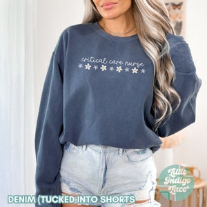 May include: A dark blue sweatshirt with the text "critical care nurse" and a row of white daisies. The sweatshirt is tucked into denim shorts.