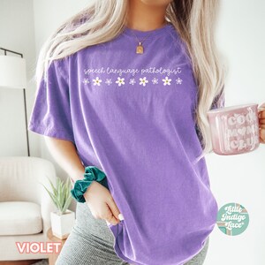 May include: Purple t-shirt with the text "speech language pathologist" and a row of white daisies with yellow centers.