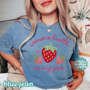 May include: A blue t-shirt featuring a graphic of three strawberries with green gingham leaves and small star accents. The text reads "women's health is my jam" in a red cursive font. The shirt has short sleeves and a crew neckline.