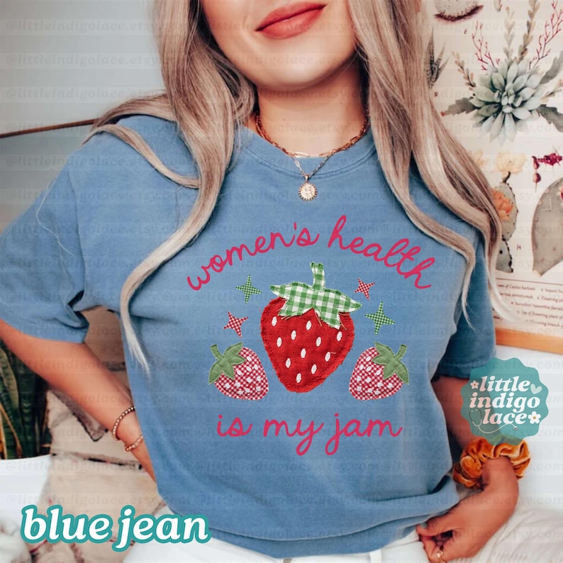 May include: A blue t-shirt featuring a graphic of three strawberries with green gingham leaves and small star accents. The text reads "women's health is my jam" in a red cursive font. The shirt has short sleeves and a crew neckline.