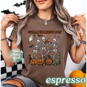 May include: Espresso-colored t-shirt featuring a Halloween design. The graphic includes dancing skeletons, pumpkins, bats, and the phrase "CAN A DEAD MAN DO IT?". A ghost and a tombstone are also part of the design.