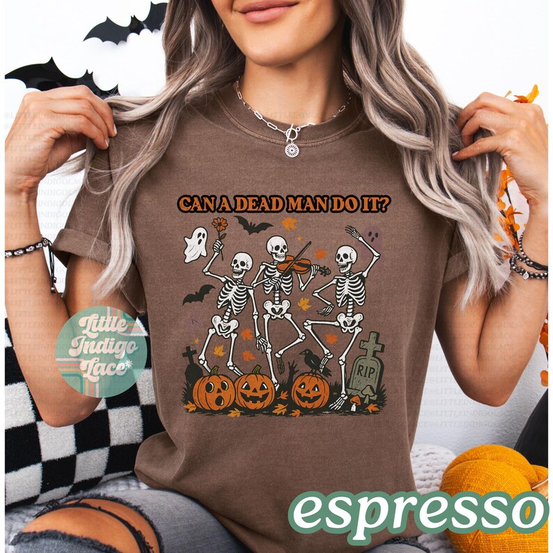 May include: Espresso-colored t-shirt featuring a Halloween design. The graphic includes dancing skeletons, pumpkins, bats, and the phrase "CAN A DEAD MAN DO IT?". A ghost and a tombstone are also part of the design.