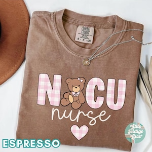 Puede incluir: Una camiseta marr&oacute;n con el texto "NICU nurse" en letras de cuadros rosas y blancos. Un coraz&oacute;n de cuadros rosas y blancos est&aacute; debajo del texto. Un oso de peluche marr&oacute;n con un lazo rosa est&aacute; en el medio del texto. El texto "ESPRESSO" est&aacute; en la parte inferior de la camiseta.
