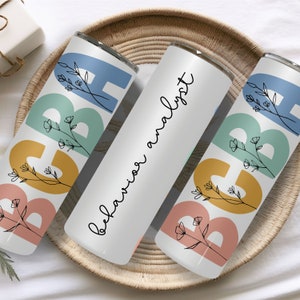 May include: Three white insulated tumblers with colorful designs. Two tumblers feature the letters "ABA" in blue, green, yellow, and pink, with floral accents. The center tumbler reads "Behavior Analyst" in black script. All tumblers are on a woven tray.