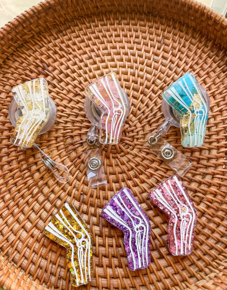 Pu&ograve; includere: Sei portachiavi in resina colorati a forma di osso del ginocchio umano con glitter e scaglie dorate. I portachiavi sono su una superficie di vimini intrecciato.