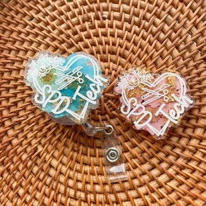 May include: Two heart-shaped resin keychains with a floral design and the text "Spa Tech" in white lettering. The keychains are attached to a clear retractable badge holder.