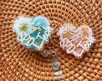 May include: Two heart-shaped resin keychains with a floral design and the text "Spa Tech" in white lettering. The keychains are attached to a clear retractable badge holder.