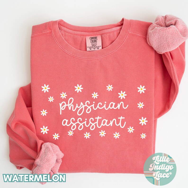 May include: A pink sweatshirt with the text "physician assistant" in white cursive lettering surrounded by white daisies. The sweatshirt is folded in half and the brand label is visible.