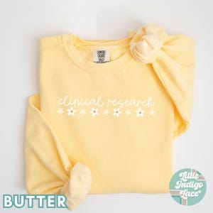 May include: A yellow sweatshirt with the text "clinical research" in white script with a row of white daisies below. The sweatshirt is folded and the sleeves are rolled up.