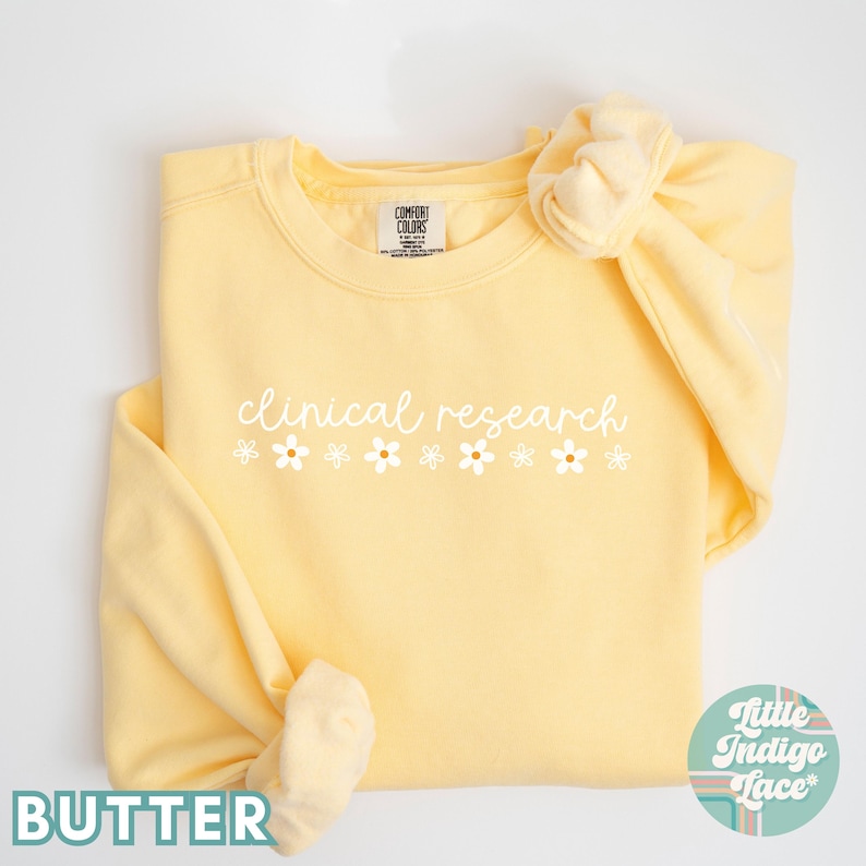 May include: A yellow sweatshirt with the text "clinical research" in white script with a row of white daisies below. The sweatshirt is folded and the sleeves are rolled up.