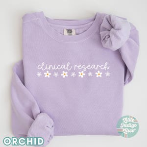 May include: A lavender sweatshirt with the text "clinical research" in white script with a row of white daisies with yellow centers below the text.