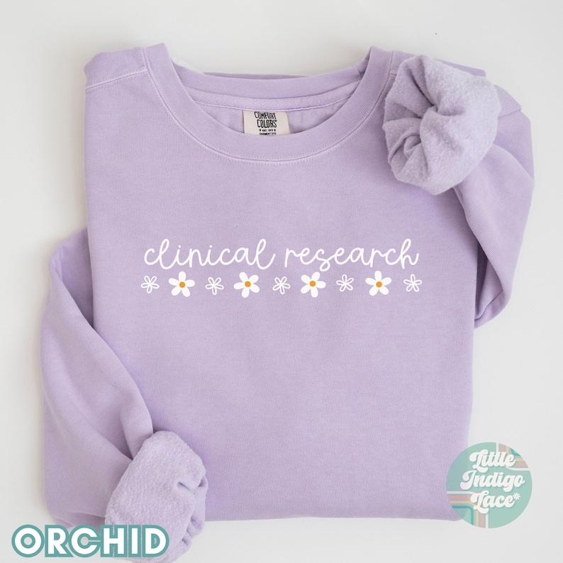 May include: A lavender sweatshirt with the text "clinical research" in white script with a row of white daisies with yellow centers below the text.