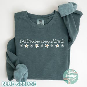 May include: Blue spruce-colored sweatshirt with the words "lactation consultant" in white script, accompanied by a row of white daisies with yellow centers. The text "BLUE SPRUCE" is at the bottom. The sweatshirt has a crew neck and long sleeves.
