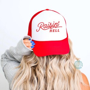 May include: A red and white trucker cap featuring the text Raisin' HELL in red script and block lettering on the front panel. The cap has a solid red brim and a white front panel with a red mesh back, designed with a structured crown and adjustable fit.