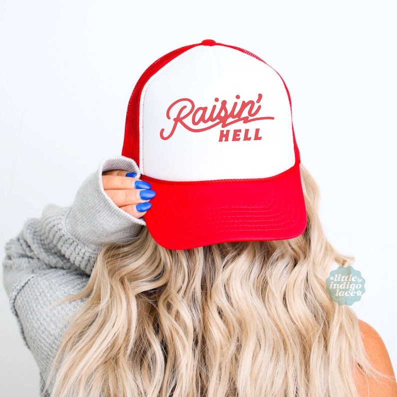 May include: A red and white trucker cap featuring the text Raisin' HELL in red script and block lettering on the front panel. The cap has a solid red brim and a white front panel with a red mesh back, designed with a structured crown and adjustable fit.
