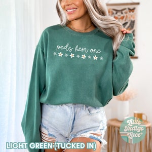 May include: A light green sweatshirt with the text "peds hem onc" and a row of white daisies printed on the front. The sweatshirt is tucked into light blue denim shorts.