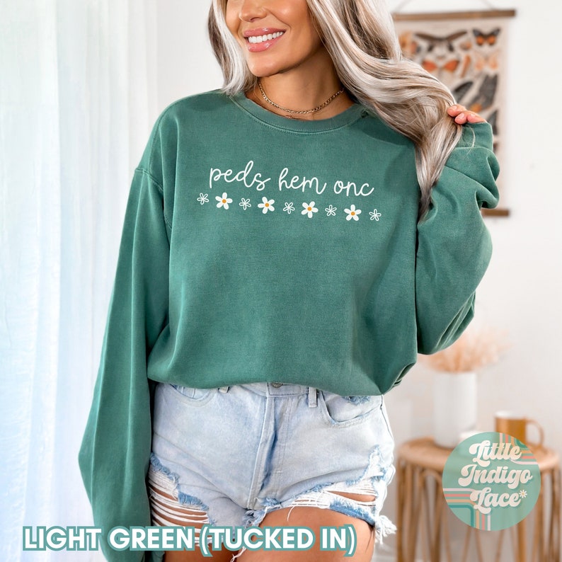 May include: A light green sweatshirt with the text "peds hem onc" and a row of white daisies printed on the front. The sweatshirt is tucked into light blue denim shorts.