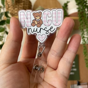 May include: A pink and white checkered badge holder with the text "NICU nurse" and a small brown teddy bear with a pink bow. The badge holder has a clear plastic clip.