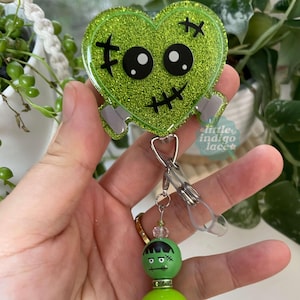 May include: A heart-shaped badge reel featuring a bright green glitter finish with a black stitched face design and two grey bolts. Attached is a clear plastic strap and a beaded charm featuring a green character head with black hair and stitches.