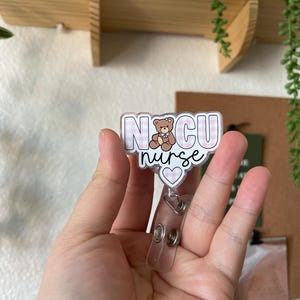 May include: A pink and white checkered badge holder with the text "NICU nurse" and a small brown teddy bear wearing a pink bow. The badge holder has a clear plastic retractable badge reel.