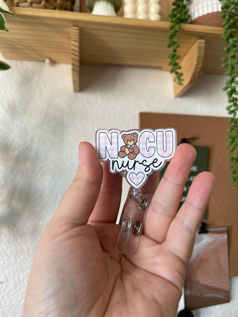 May include: A pink and white checkered badge holder with the text "NICU nurse" and a small brown teddy bear wearing a pink bow. The badge holder has a clear plastic retractable badge reel.