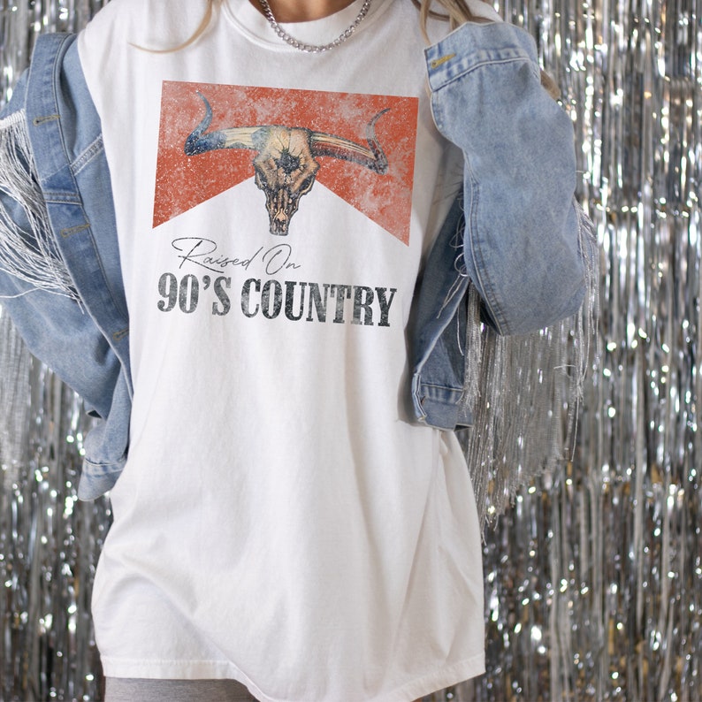 May include: White t-shirt with a distressed graphic of a bull skull and the text "Raised On 90's Country".