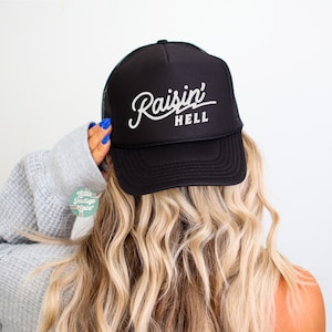 May include: A black trucker cap featuring a mesh back and a solid front panel. The front of the cap displays the white text 'Raisin' HELL' in a stylised script and block font combination. The cap has a structured crown and a curved brim.