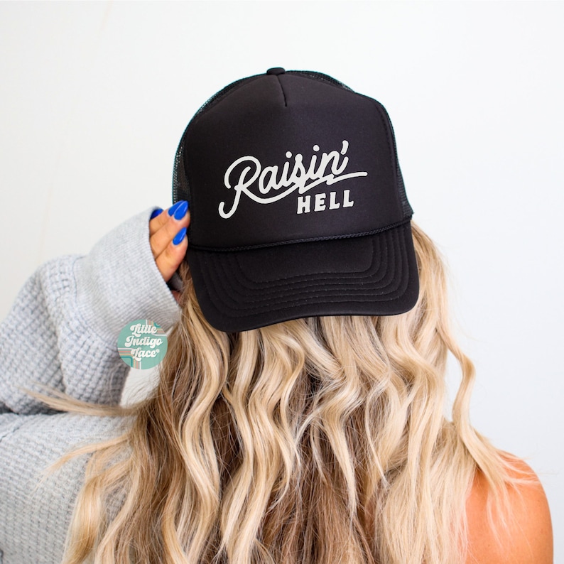 May include: A black trucker cap featuring a mesh back and a solid front panel. The front of the cap displays the white text 'Raisin' HELL' in a stylised script and block font combination. The cap has a structured crown and a curved brim.