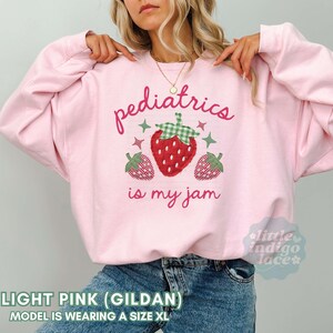 May include: A light pink crewneck sweatshirt featuring a central graphic of three strawberries with green gingham leaves and small star accents. The text reads "pediatrics is my jam" in a cursive pink font. The sweatshirt has long sleeves.