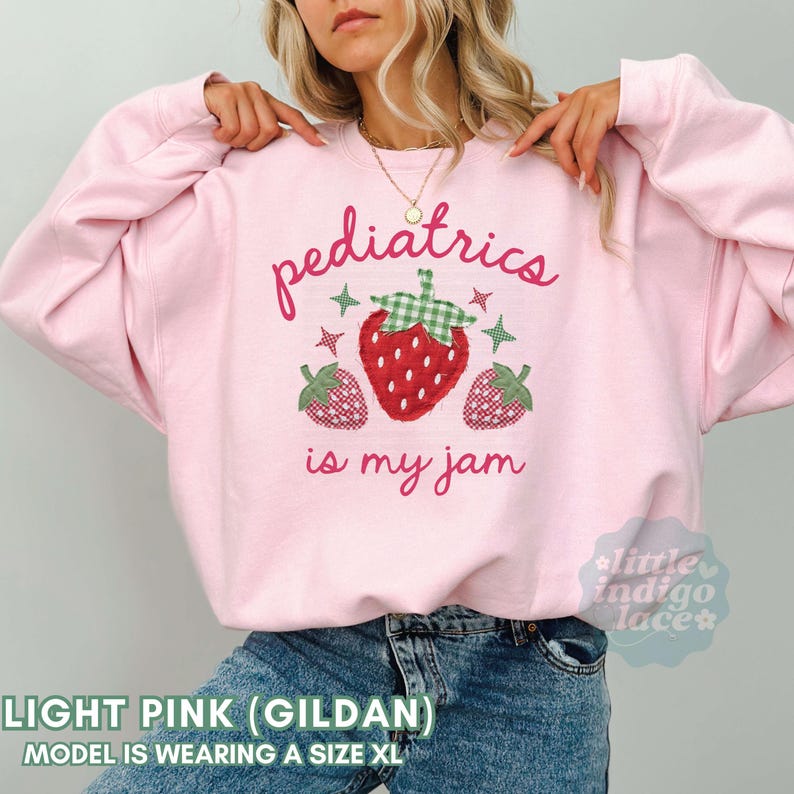 May include: A light pink crewneck sweatshirt featuring a central graphic of three strawberries with green gingham leaves and small star accents. The text reads "pediatrics is my jam" in a cursive pink font. The sweatshirt has long sleeves.