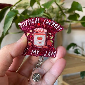 May include: A glittery red badge reel with the words "PHYSICAL THERAPY IS MY JAM" in white. The badge features a jar of strawberry jam with two strawberries. The reel has a clear plastic strap and a metal clip.