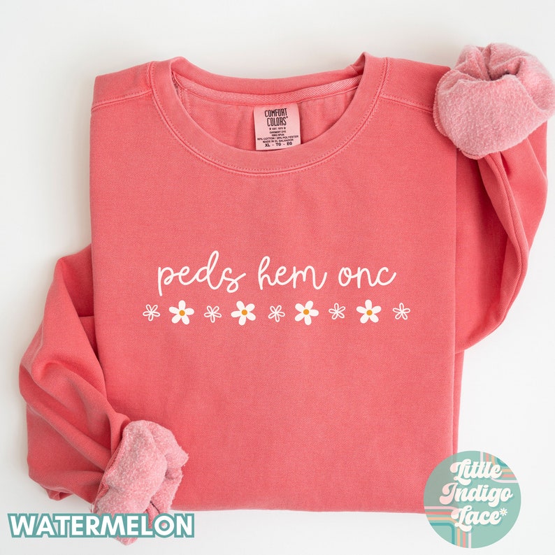 May include: A coral pink sweatshirt with the text "peds hem onc" in white script and a row of white daisy flowers below the text. The sweatshirt is folded in half and the sleeves are rolled up.