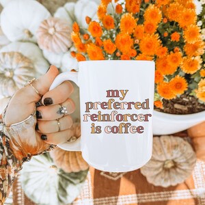 May include: White ceramic coffee mug with the text "my preferred reinforcer is coffee" in brown, pink, and orange lettering. The mug is being held by a hand wearing a brown and orange patterned glove.