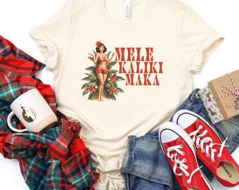 May include: A cream-colored t-shirt featuring a graphic of a pin-up style figure standing in front of a Christmas tree with the red text Mele Kaliki Maka. The shirt is displayed with a red and green plaid fabric, a white mug, blue jeans, and red sneakers.