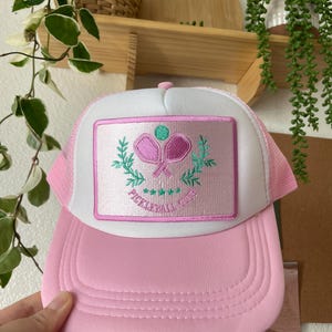 May include: A pink and white trucker hat with a pink and green embroidered patch that says "Pickleball Club".